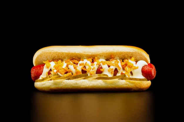Photo appetizing hot dog with grilled beef sausage , pickles, mayonnaise and fried onions on black