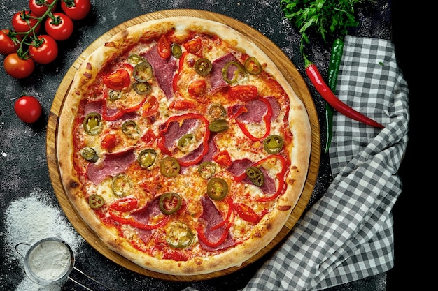 Appetizing italian pizza with ham, cheese, jalapenos, tomatoes and bell pepper in a composition with ingredients on a black table