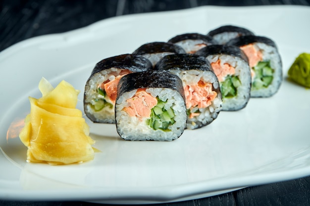 Appetizing Japanese futomak sushi roll with salmon, cucumber in a white plate on a black wooden surface with ginger and wasabi. Japanese cuisine. Futomak roll