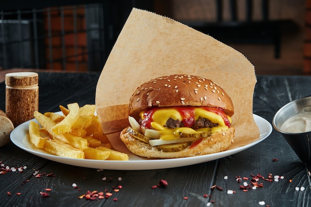 Appetizing and juicy burger with beef, melted cheddar cheese, ketchup, onions and pickled cucumbers on a white plate with french fries. Close up