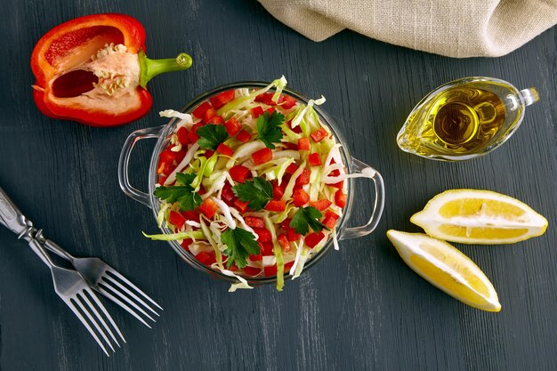 An appetizing light salad of white cabbage bell pepper and parsley dressed with a mixture of olive oil and lemon in a glass bowl surrounded by ingredients on a black wooden table