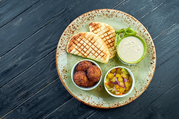 Un piatto orientale appetitoso, falafel con pane pita, servito con salsa bianca e sottaceti in un piatto verde su una superficie scura e legnosa. cibo vegetariano