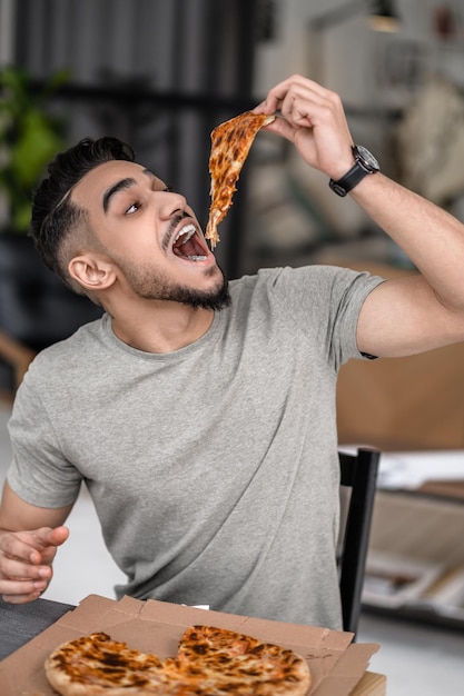 食欲をそそるピザ。若いあごひげを生やした空腹の男が家でテーブルに座って口を開けてピザの一部を保持しています。