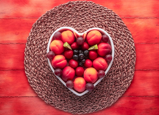 アプリコット ブドウ イチゴ ブルーベリーとキウイ木製フクシア色の背景と健康的な食事の白いハート型プレートで季節の熟した新鮮なフルーツの食欲をそそるプレート
