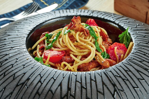 An appetizing portion of homemade Italian pasta (spaghetti) with forest mushrooms (chanterelles) cherry tomatoes and ruccola in a black plate on a wooden table. Restaurant table setting