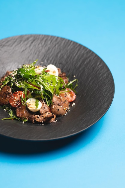 Appetizing salad with meat, egg and greens in a deep black plate on a blue background.