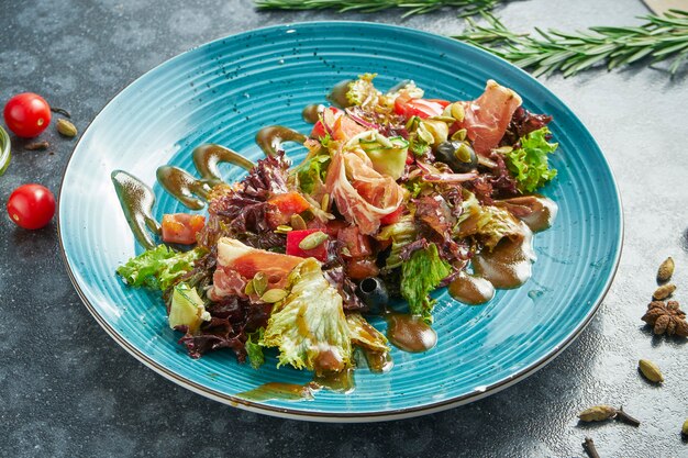 Foto insalata appetitosa con prosciutto di parma, lattuga, olive e salsa dolce su un piatto di ceramica blu su una superficie scura. vista da vicino