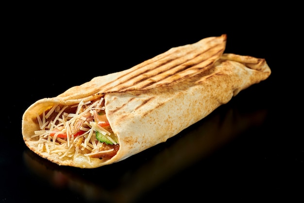 Appetizing shawarma with chicken and mozzarella, cucumbers, carrots and tomatoes in pita bread. Black surface. Isolated. Close up, selective focus