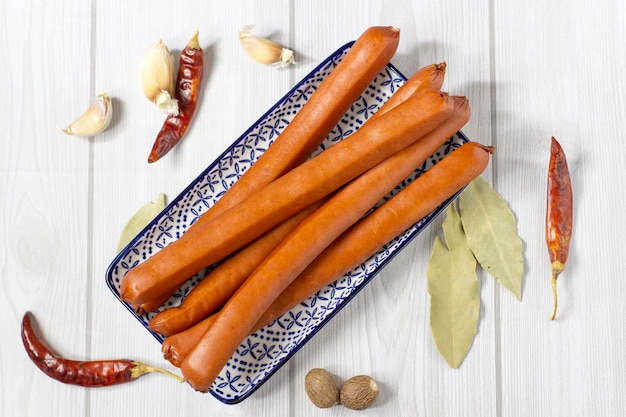 Appetizing smoked sausages on a plate with blue pattern with hot peppers on a light grey wooden background close up