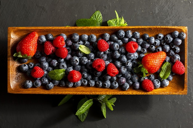 Appetizing variety of forest fruits strawberries big strawberries raspberries blueberries red berries on a wooden tray and mint leaves peppermint On textured dark surface Top view with copy s