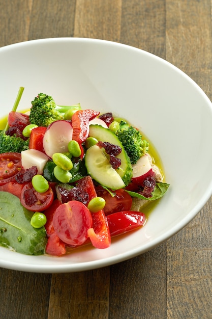 Appetizing vegetable salad with cucumber, tomatoes, radish, beans and broccoli with green butter in a white bowl on wooden table