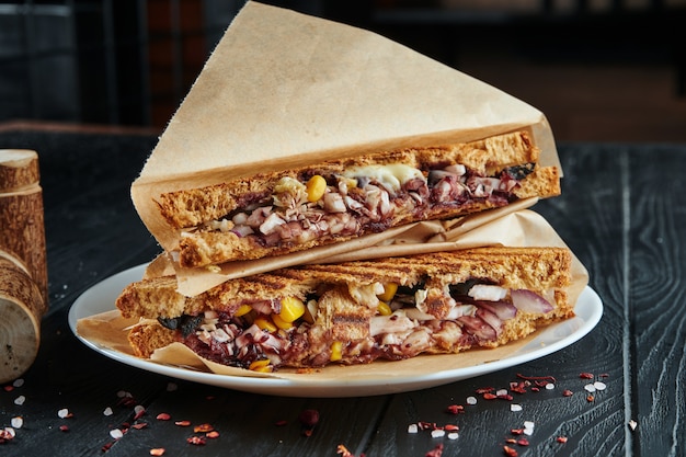 Appetizing veggie club-sandwich in toast bread with red onion, corn, cabbage and teriyaki sauce in craft paper on a black wooden. Close up