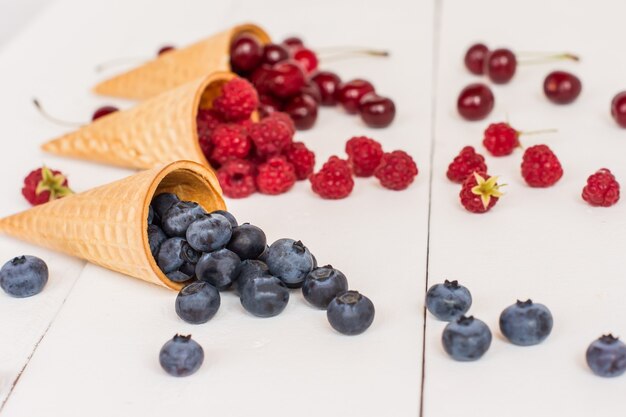 Appetizing waffle cones with blueberries, raspberries, cherries on a white wooden background. Summer dessert.