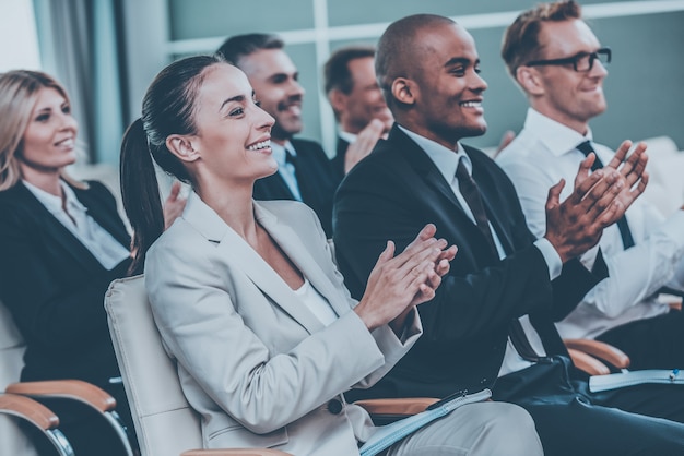 Applauding to speaker. Group of happy business people in formalwear sitting at the chairs in conference hall and applauding