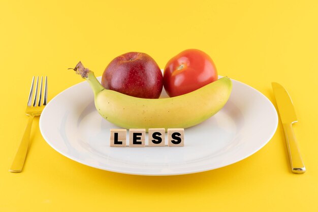 Photo apple a banana and a tomato on a plate and the word less written on wooden blocks