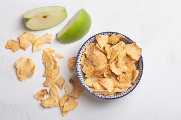 Apple chips in a white bowl and pieces of this fruits on a table