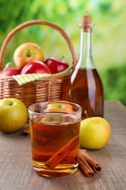 Apple cider in glass and bottle with cinnamon sticks and fresh apples on wooden table on bright background