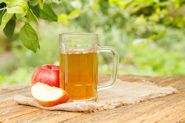 Photo apple cider in glass goblet and fresh red apples on wooden boards with blurred green natural background in daylight. shellow depth of field. organic drinks