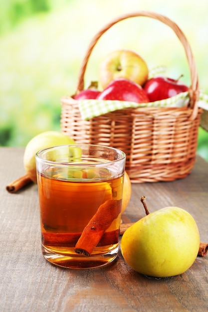 Apple cider in glass with cinnamon sticks and fresh apples in wicker basket on wooden table on bright background