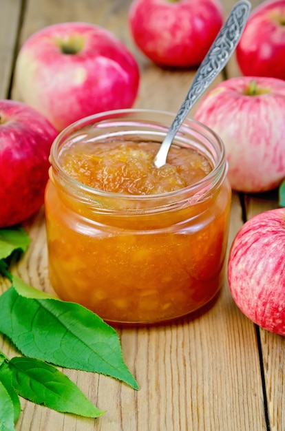 Foto marmellata di mele in un barattolo di vetro con un cucchiaio, mele rosse fresche, ramoscelli con foglie su una tavola di legno