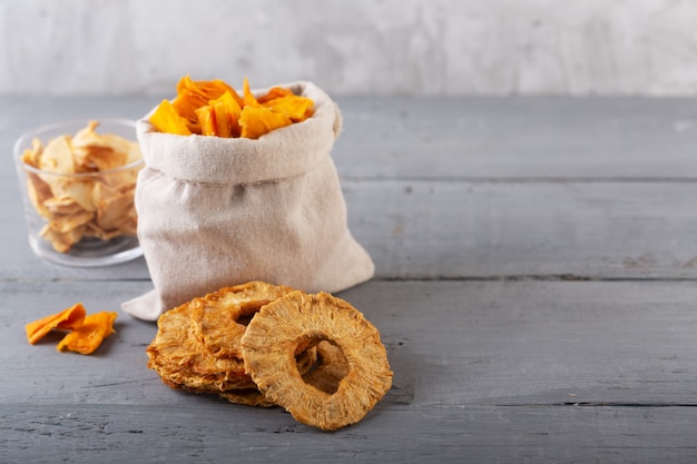 Apple, pineapple and mango chips in a jar and pieces of this fruits on a table