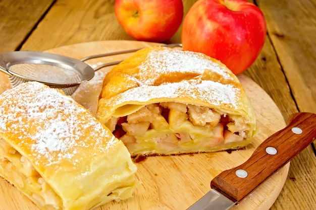 Foto strudel di mele con zucchero a velo, mele, colino da tè, coltello su uno sfondo di assi di legno