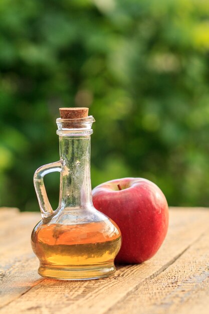 Apple vinegar in glass bottle with cork and fresh red apple on wooden boards with blurred green natural background. Organic food for health