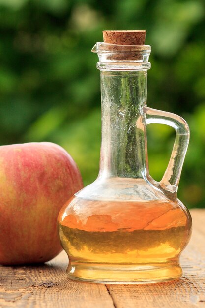 Apple vinegar in glass bottle with cork and fresh red apple on wooden boards with blurred green natural background. Organic food for health