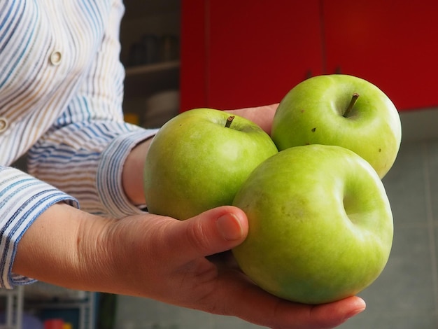 Foto mele in mano la donna tiene in mano tre mele verdi e le offre allo spettatore alimenti biologici frutta fresca per il cibo grande mela acida camicia bianca a righe cucina con mobili rossi