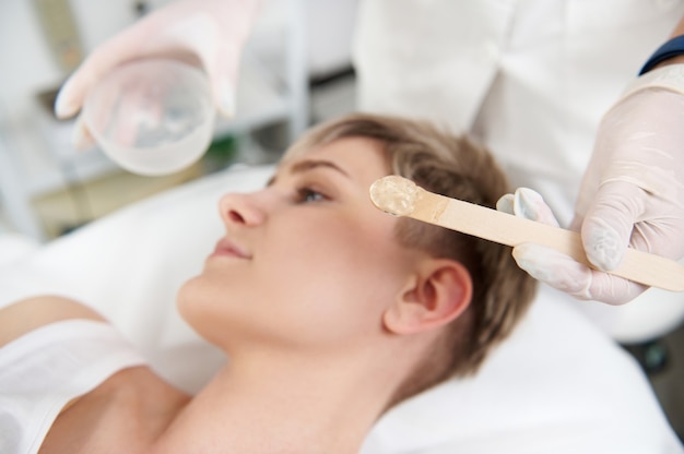 Appliance of transparent anaesthetic gel on woman's face for making the laser removal hair procedure non painful. Professional laser treatment in modern SPA clinic. Focus on wooden spatula