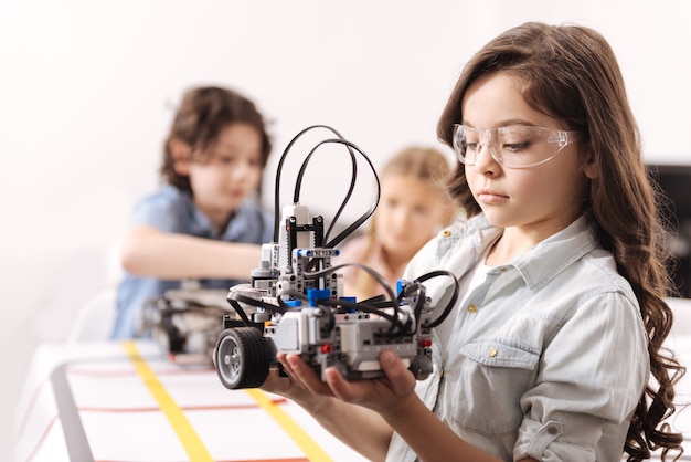 Applying my knowledge. Involved skilled smart girl standing at school and holding robot while colleagues working on the project