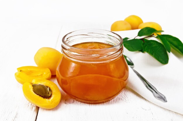 Apricot jam in glass jar, fruits, branch with leaves, a napkin and a spoon on the background of light wooden board