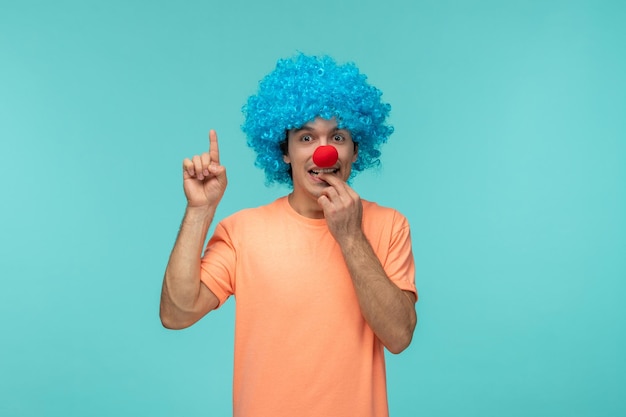 Photo april fools day guy clown awkward pointing finger upbiting nail blue hair excited funny red nose