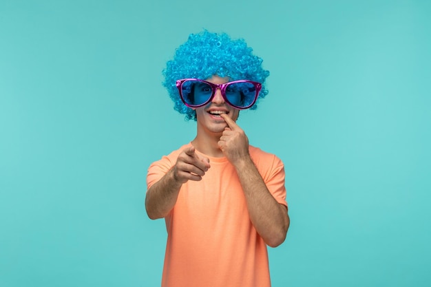 Foto pesce d'aprile ragazzo pagliaccio divertente capelli blu blu rosa grandi occhiali da sole che si mordono le dita che puntano davanti