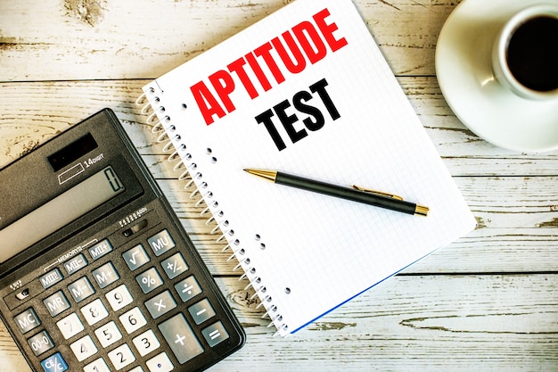 APTITUDE TEST geschreven op wit papier in de buurt van koffie en rekenmachine op een lichte houten tafel