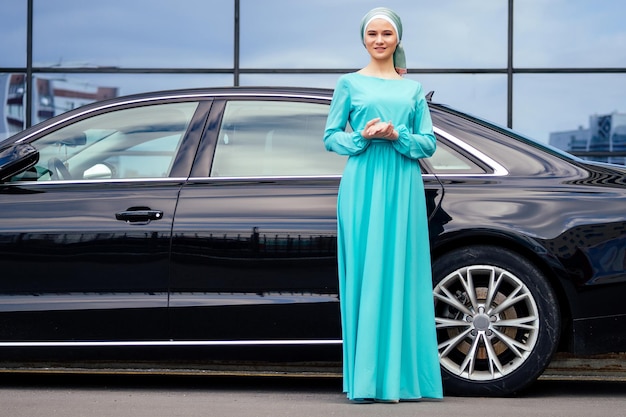 Arab business woman hijab standing in front of her luxury car on the street on a background of skyscrapers of Dubai The woman is dressed in a stylish abaya