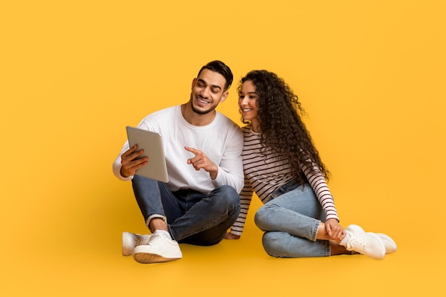 Arab Couple With Digital Tablet Relaxing On Floor Over Yellow Studio Background