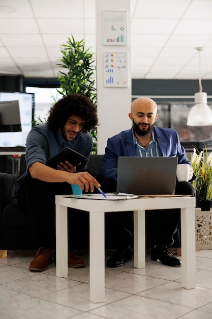 Arab coworkers analyzing financial data and creating presentation on laptop together. Company employee doing marketing analysis and planning strategy while sitting on couch