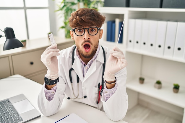 Arab doctor man with beard holding covid test in shock face looking skeptical and sarcastic surprised with open mouth