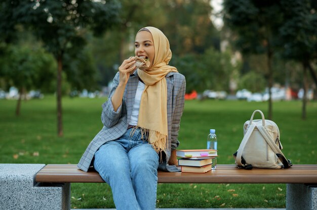 Арабская студентка, сидящая на скамейке в летнем парке. Мусульманская женщина отдыхает на пешеходной дорожке.
