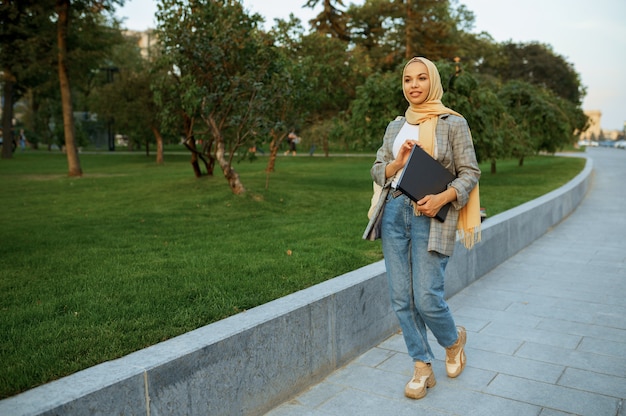 夏の公園を歩いているラップトップを持つアラブの女子学生。散歩道で休んでいるイスラム教徒の女性。