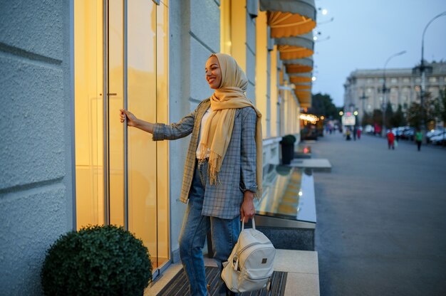 Arab girl in hijab at the entrance of fashion store in downtown. Muslim woman walking on street.