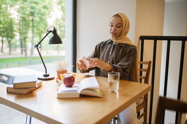アラブの女の子が大学のカフェ、上面図でコーヒーとカップの写真を作成します。図書館に座っている本を持つイスラム教徒の女性。
