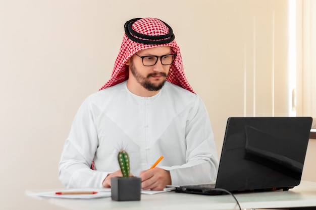 An Arab man, a businessman, a sheikh in a national Arab costume works at a table in the office. Investments, business, work via the Internet, online contracts.