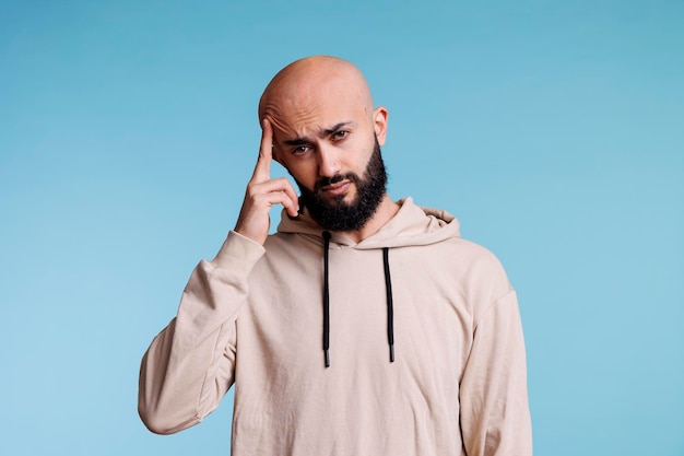 Arab man feeling pain of migraine while looking at camera with tired facial expression. Person rubbing temple while suffering from headache studio portrait on blue background