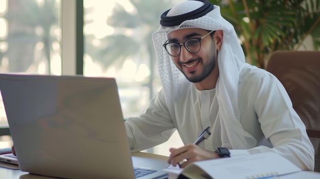 An Arab man in glasses is seated at a desk writing in a notebook while browsing the internet watching a webinar on a computer studying online and looking at the screen