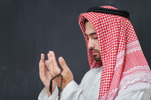 Arab man in traditional clothes praying to God or making dua. Muslim boy keeps hands in praying position in front of black chalkboard representing Ramadan concept