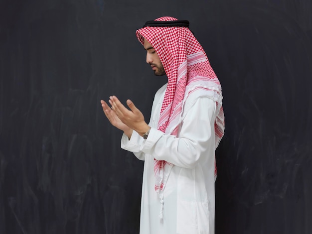 신에게 기도하거나 두아를 만드는 전통 옷을 입은 아랍 남자. 무슬림 소년은 라마단 개념을 나타내는 검은 칠판 앞에서 손을 기도 자세로 유지합니다.