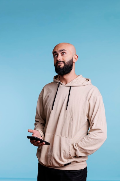 Arab man using smartphone, thinking while typing online message and looking upwards. Young person with pensive facial expression holding mobile phone while standing with hand in hoodie pocket