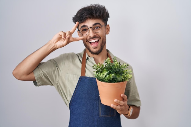 Arab man with beard holding green plant pot doing peace symbol with fingers over face, smiling cheerful showing victory